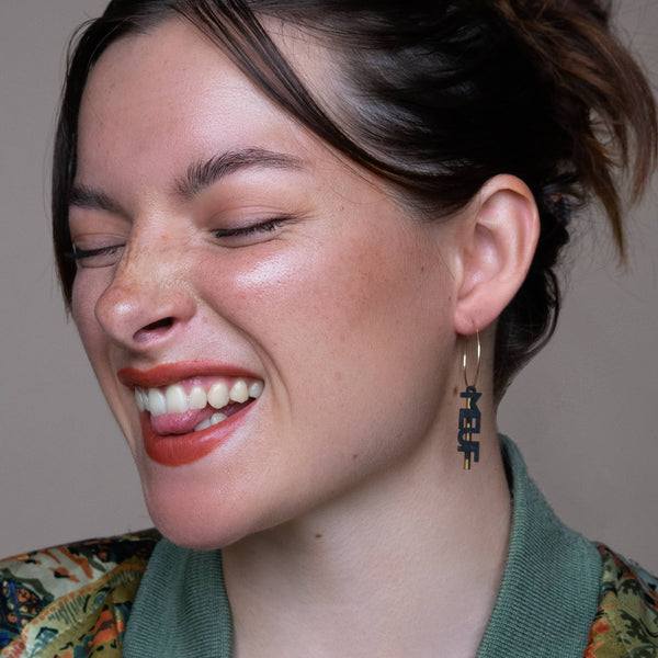 Boucles d'oreilles dépareillées Meuf en Or portées : créole argent pendentif texte MEUF noir sur modèle souriante, style rock chic