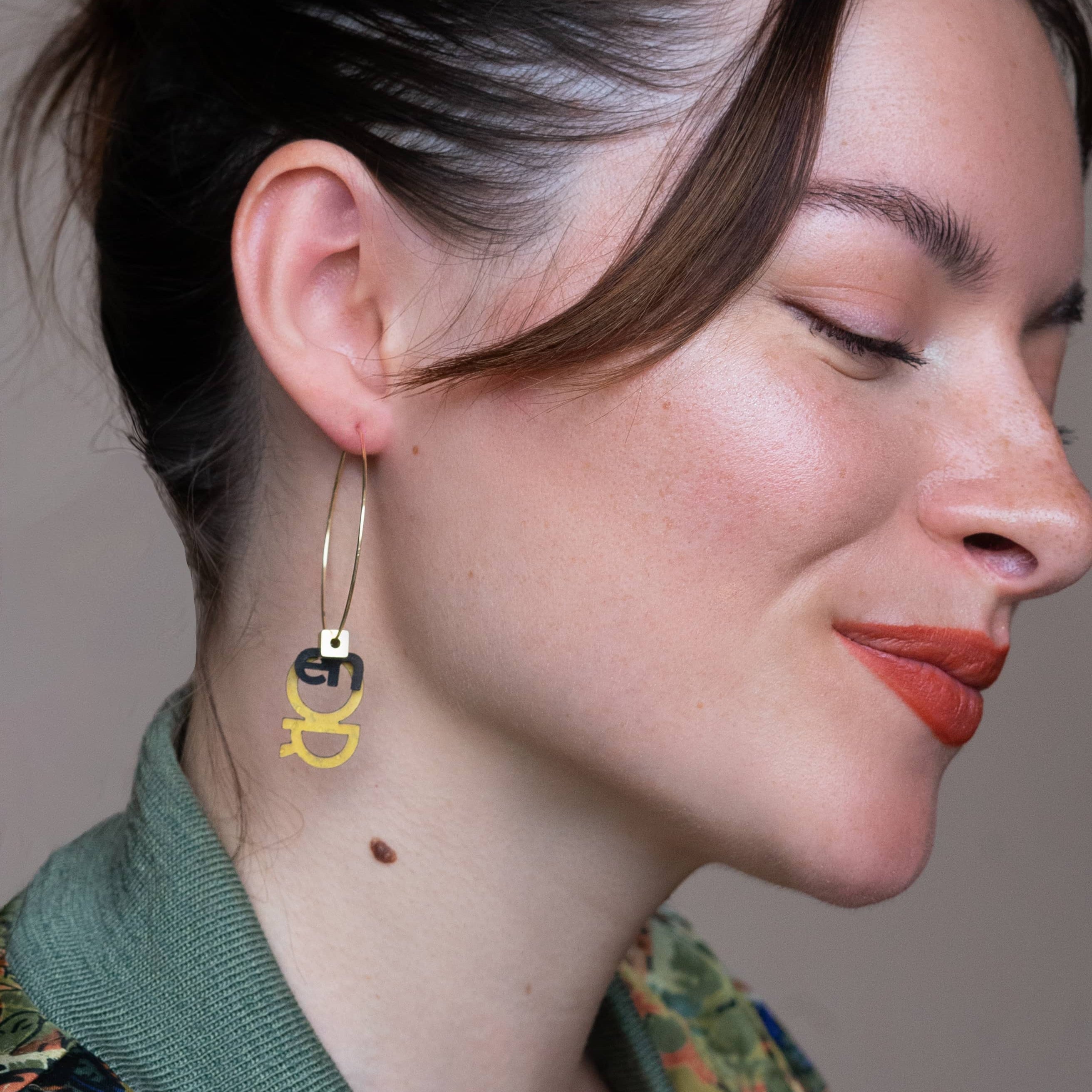Boucles d'oreilles dépareillées Meuf en Or portées de profil : créole dorée fine avec pendentifs lettres OR noir et doré