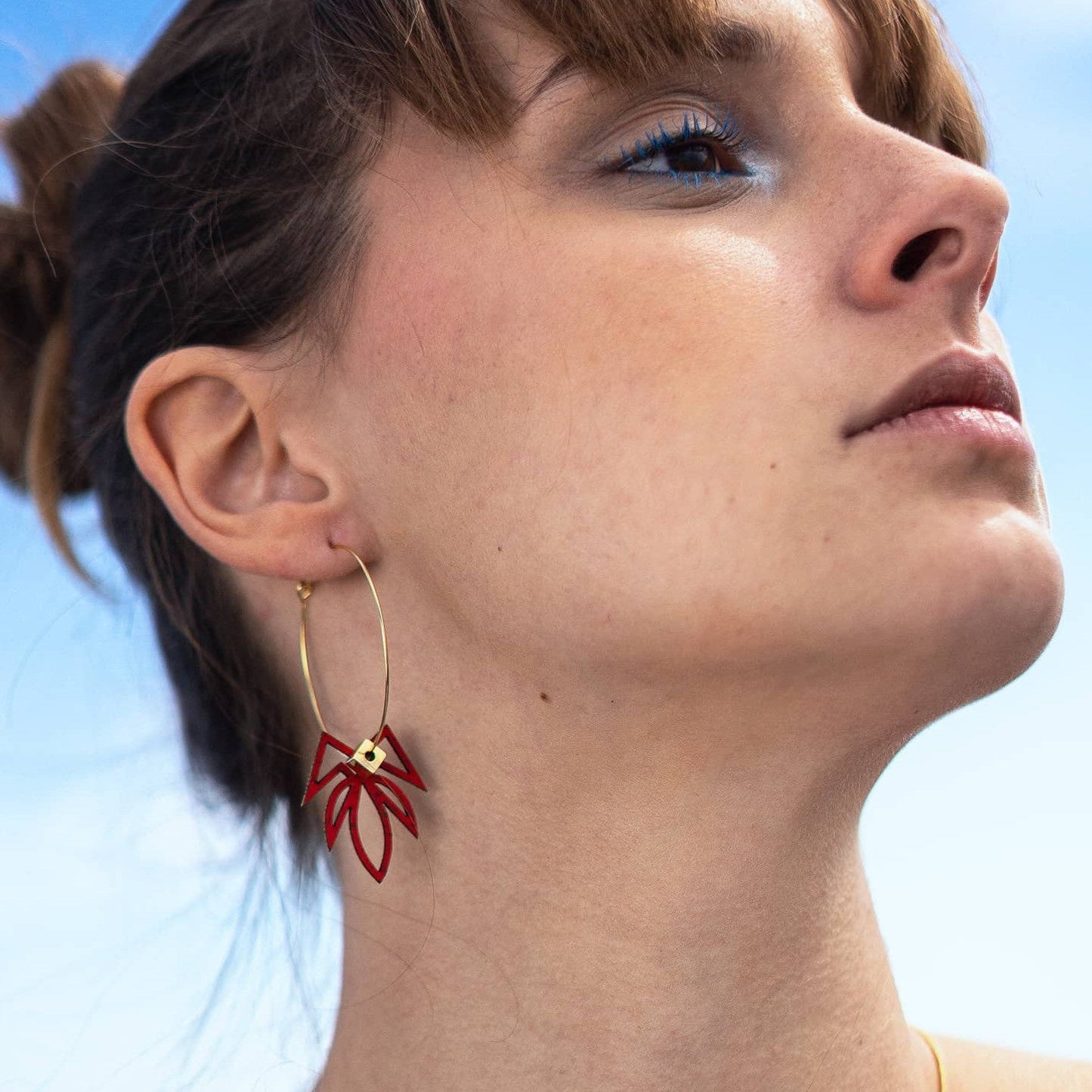 femme portant boucles d’oreilles dépareillées rouges créoles asymétriques en acier inoxydable breloque graphique rouge look moderne ciel bleu