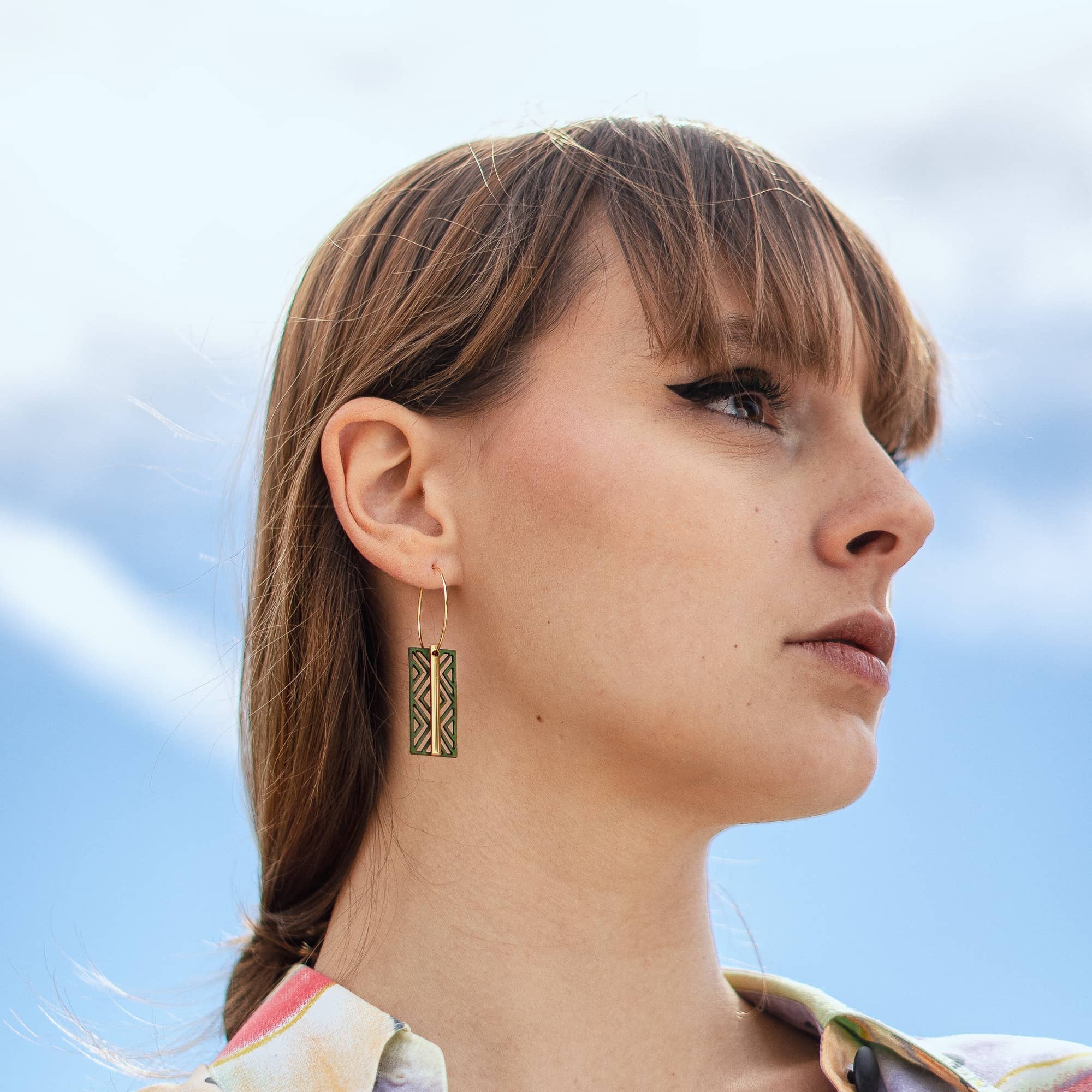 femme portant boucles d’oreilles dépareillées vertes créoles asymétriques en acier inoxydable pendentif graphique vert motif géométrique style moderne ciel nuageux