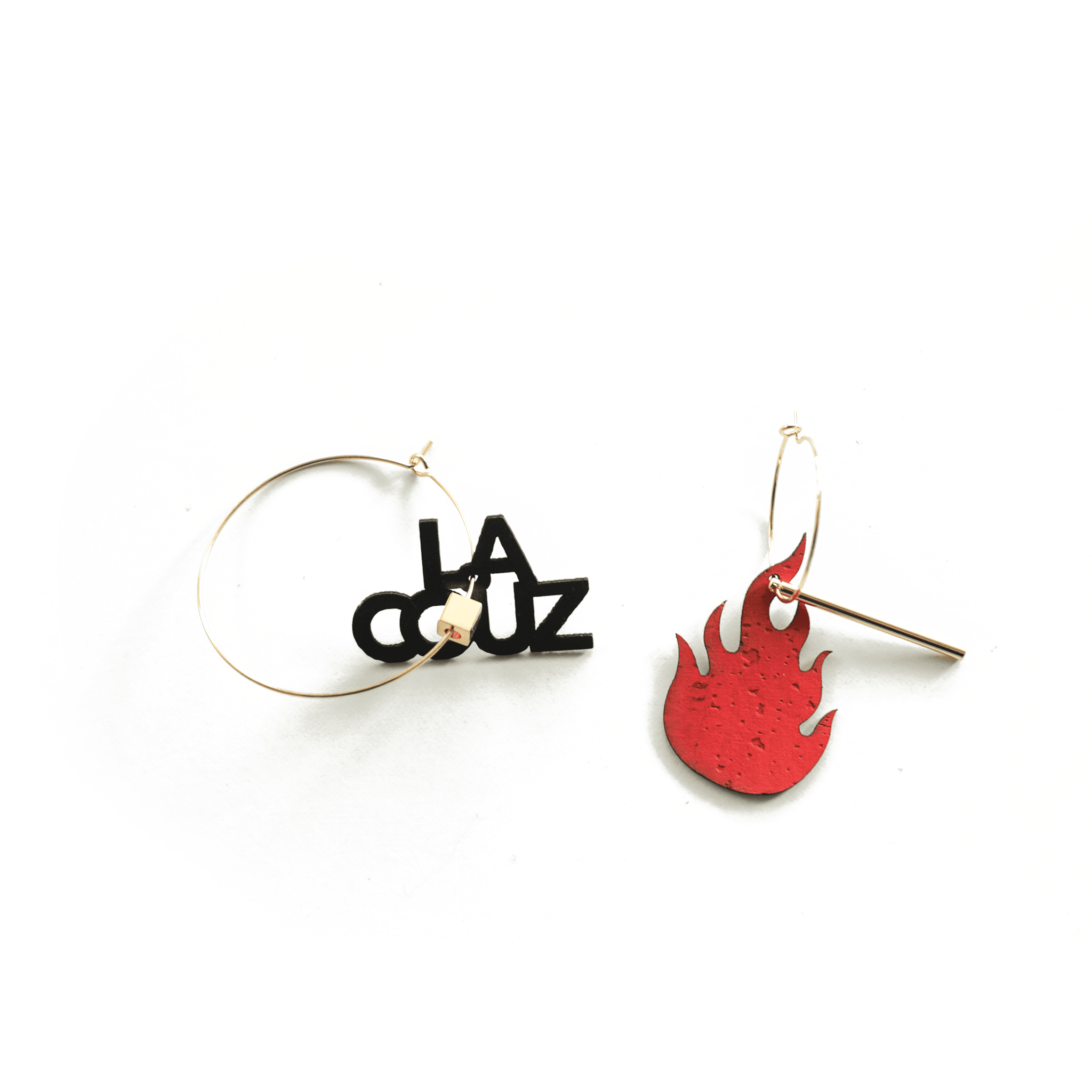 Paire de créoles dorées asymétriques : l’une avec le pendentif noir “LA COUZ” et un petit cube beige, l’autre avec une flamme rouge et une tige dorée, sur fond blanc.