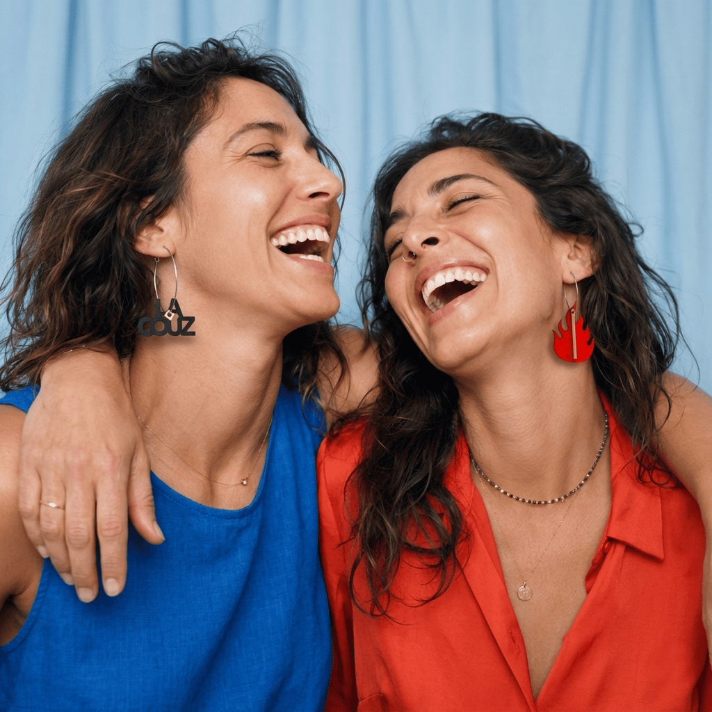 Paire de créoles dorées asymétriques : l’une avec le pendentif noir “LA COUZ” et un petit cube beige, l’autre avec une flamme rouge et une tige dorée, sur fond blanc. cadeau idéal pour cousine